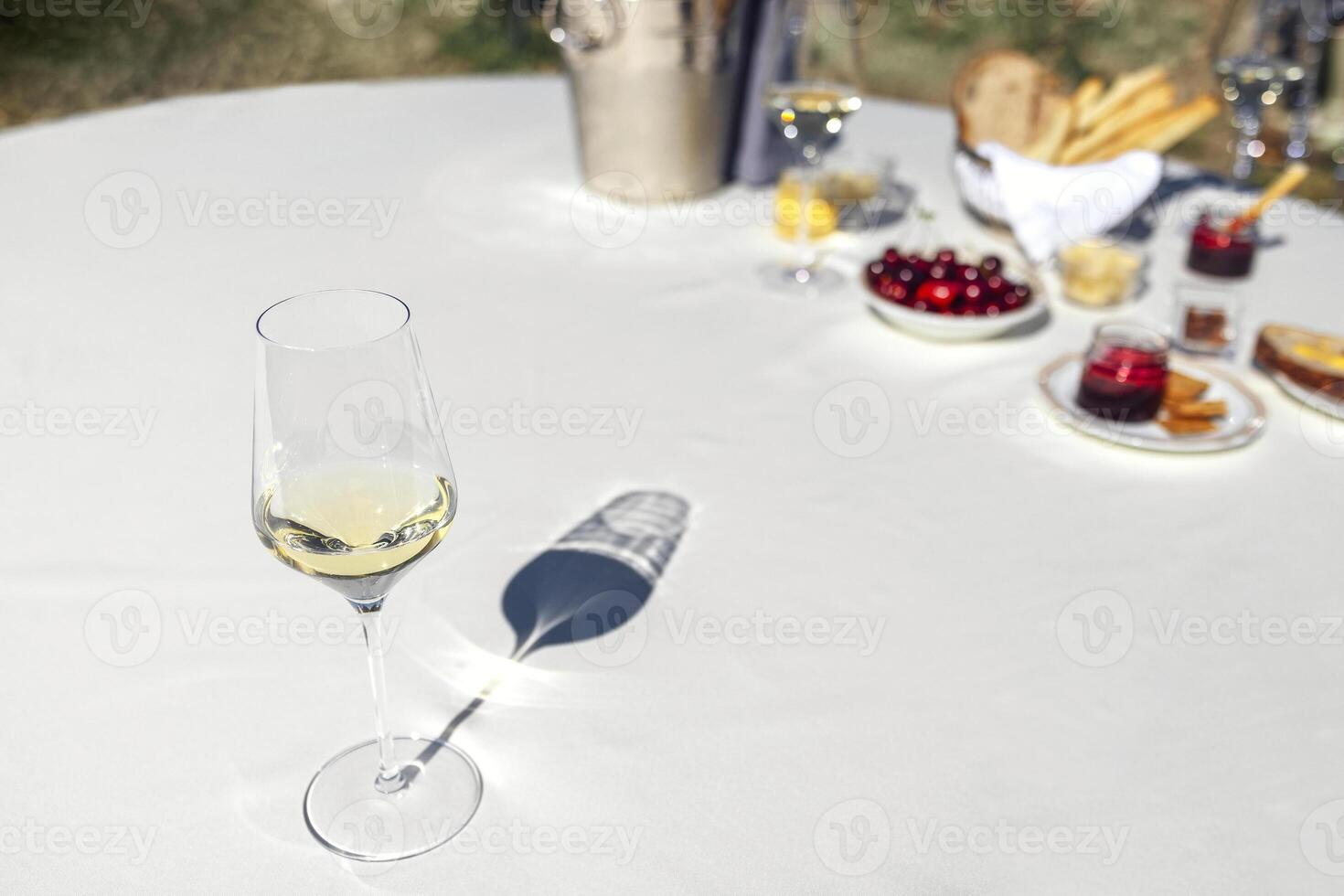 Elegant glass of delicious sparkling wine on background of table with white tablecloth and snacks. Berries, bread, cheese, ice cooler and several goblets on backdrop. Sunny day and long shadows. photo