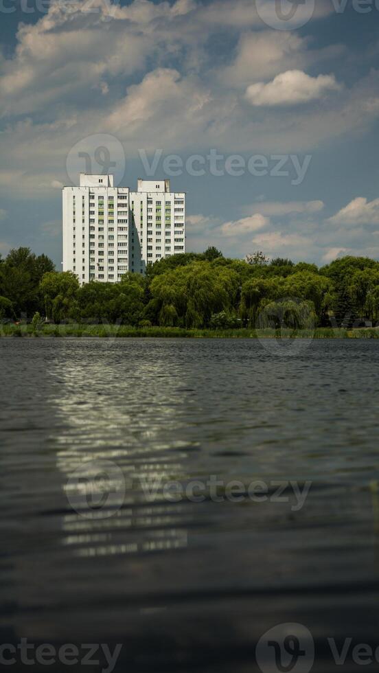 Two houses, multi-storey buildings, thick trees in front of them, and a large pond photo