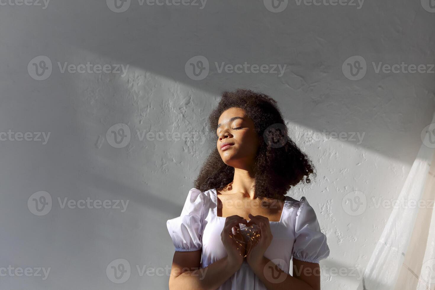 Young peaceful grateful thankful african american woman with closed eyes in white dress making heart sign, led lights in hands of tranquil black female showing symbol of love photo