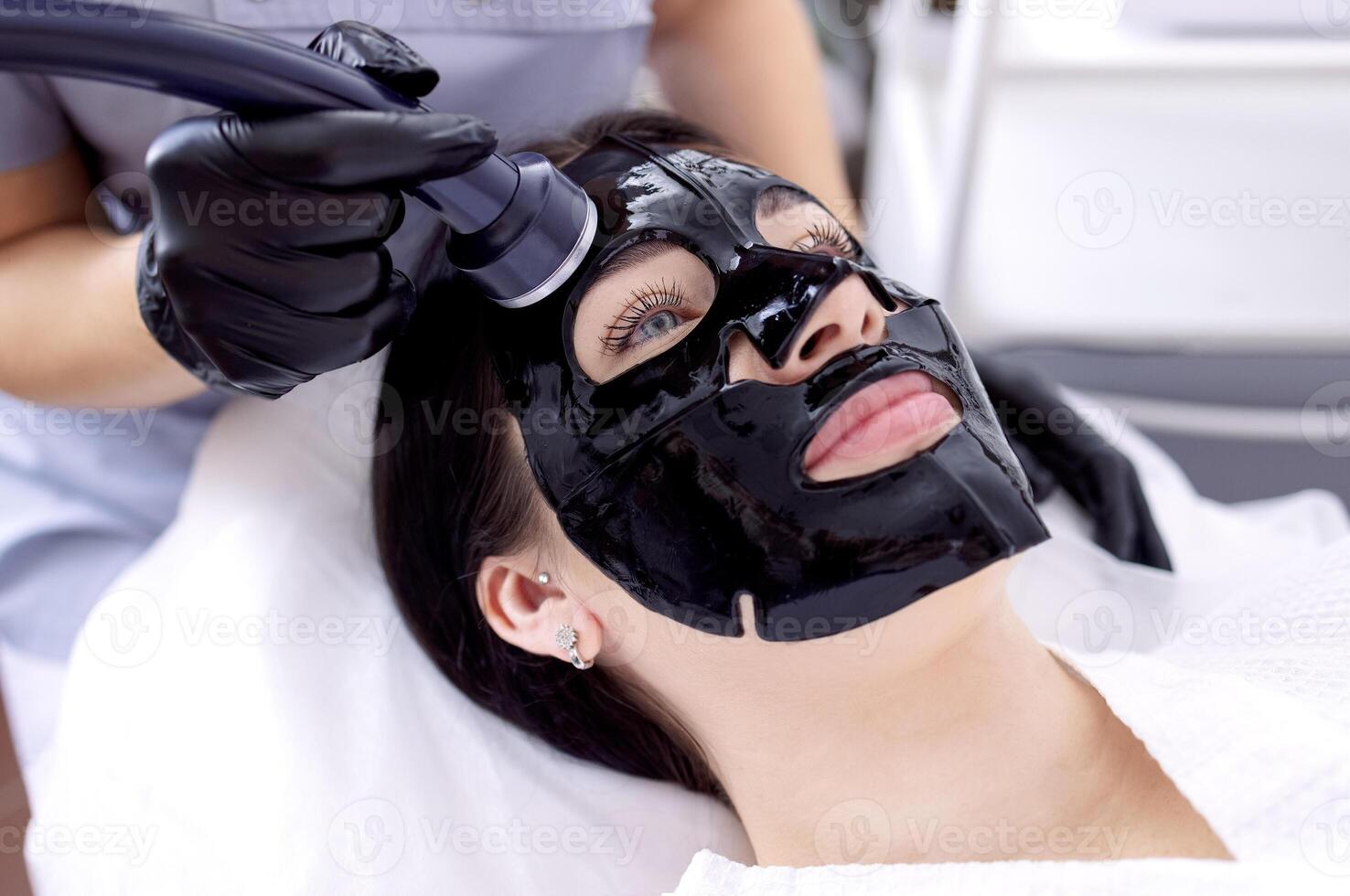 Close-up of a young woman in a black cosmetic mask at a spa procedure. A cosmetologist performs a cosmetic procedure using an ultrasound device. photo