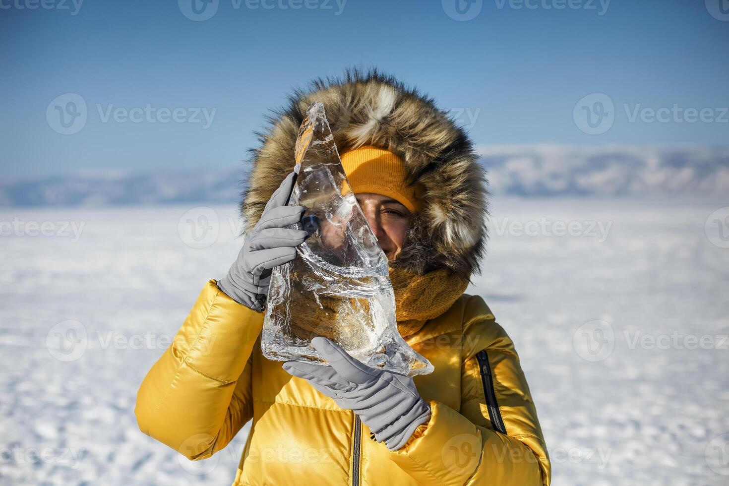 Happy woman wearing winter yellow jacket holding piece of transparent crushed ice cubes over her face photo