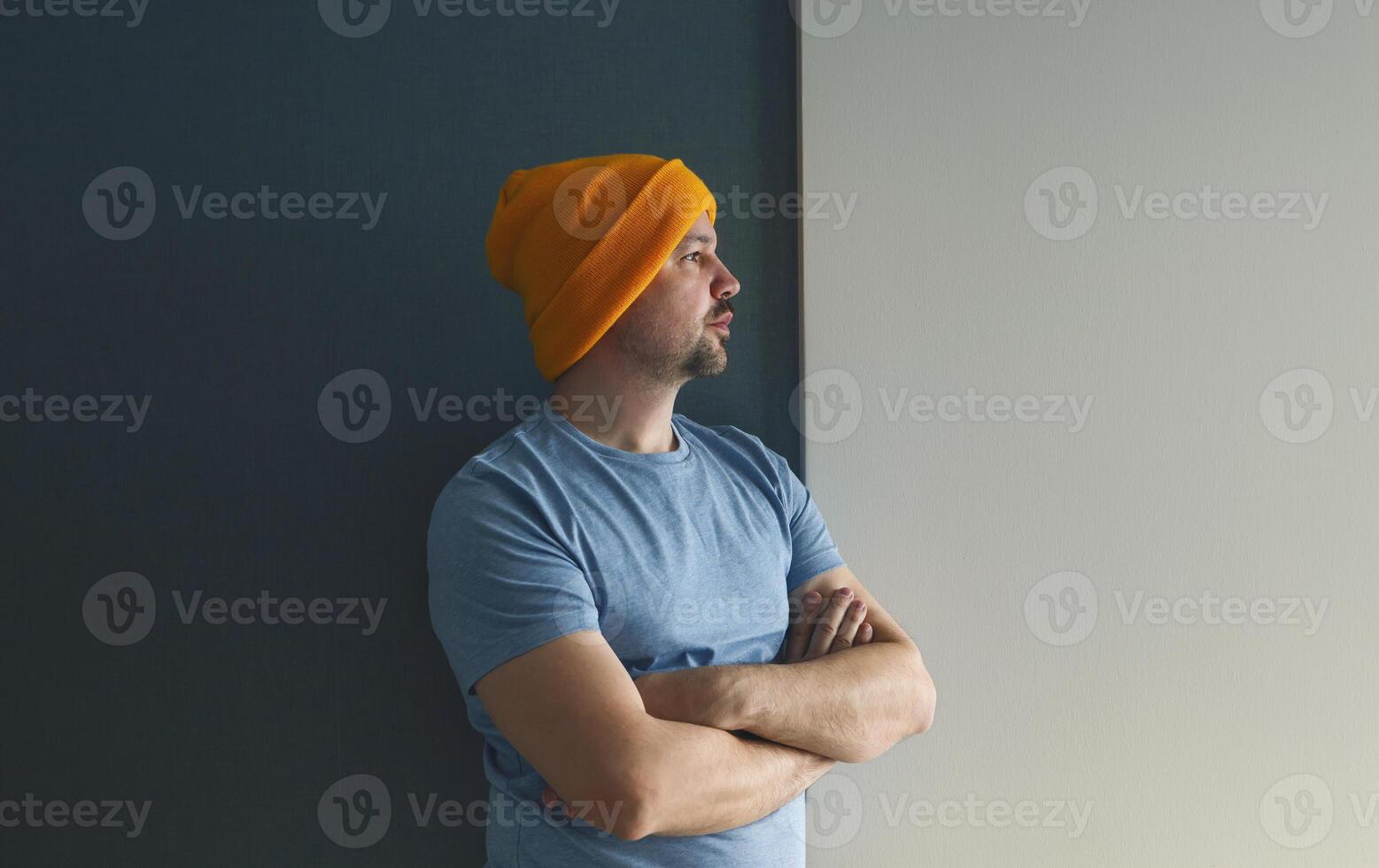 Young man in a blue T-shirt and a yellow hat stands with his arms crossed against a gray-blue wall. photo