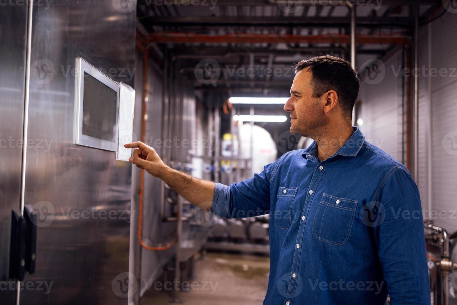 Young man sets necessary parameters on display on equipment in cheese factory. Smiling worker near milk and butter production machine. photo