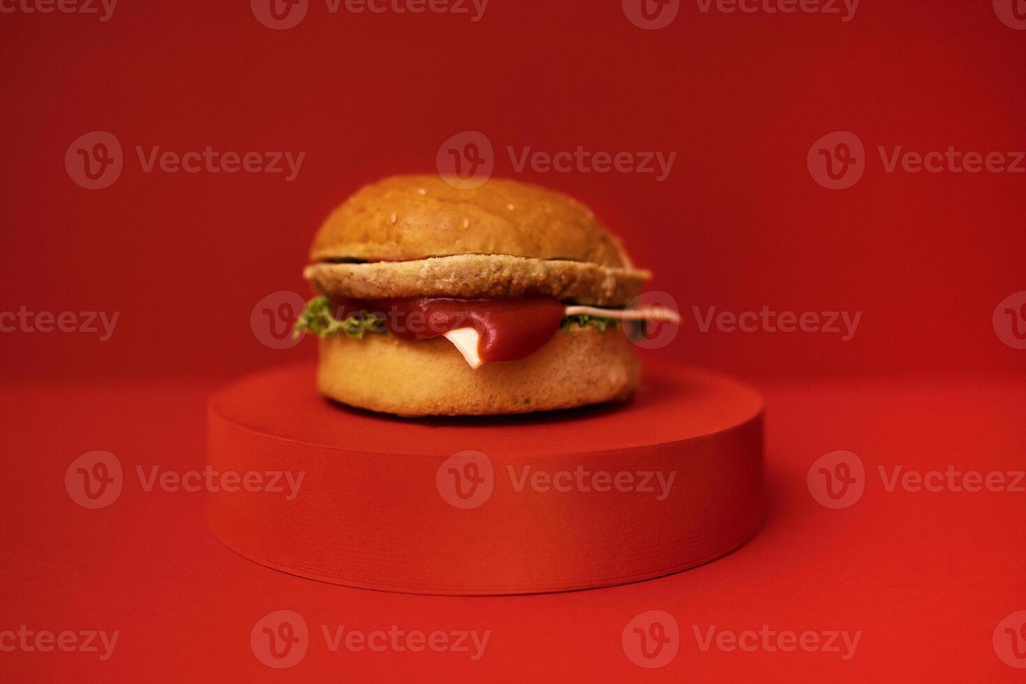 Close up of big yummy burger. Delicious sandwich with cheese, ketchup, lettuce and cutlet is on a red round stage. Red bright background. photo