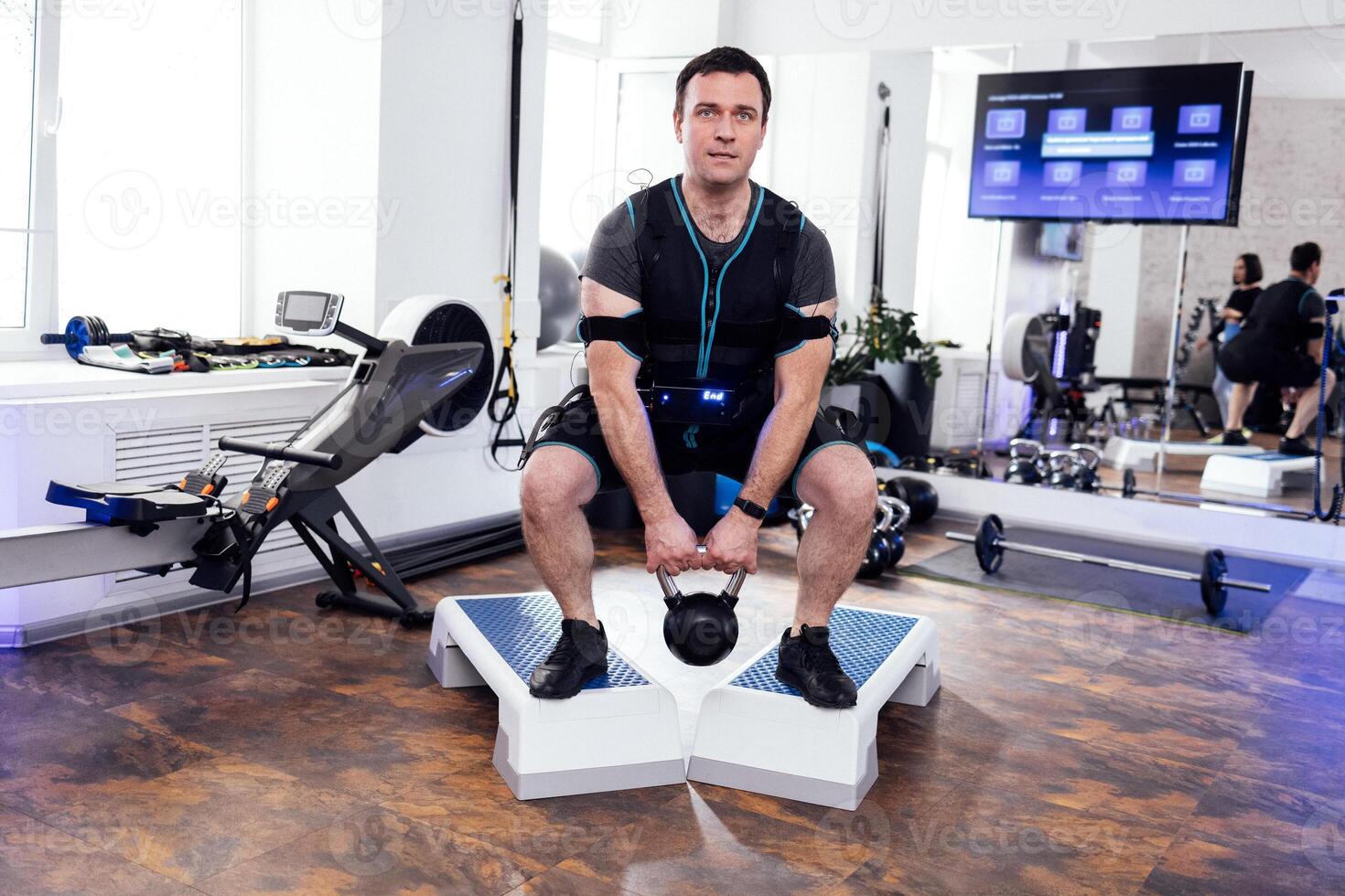 Strained man in ems suit standing on step platforms and doing squats with kettlebell. photo