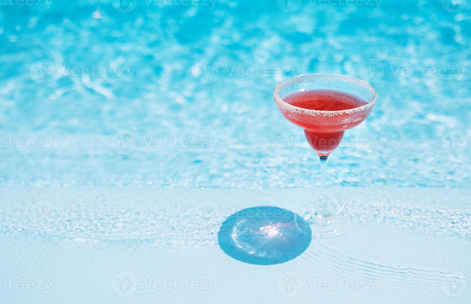 An elegant glass with long stem with a red cocktail. Tasty strawberry drink by the pool. Delicious fresh margarita. Sunny day and reflection of rays in blue water in the background. photo