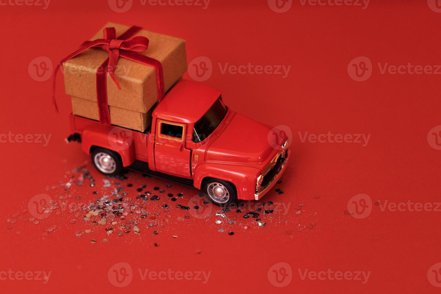 Red toy truck with craft gift box and red bow in its trunk. Miniature car with Christmas decoration on red bright background with sparkles and confetti. photo