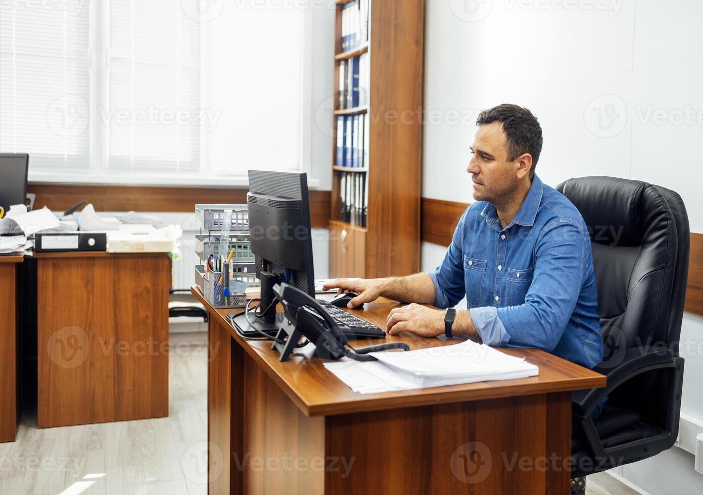 Young man in denim shirt is sitting at desk and working at personal computer in office. Worker does his job at workplace. photo