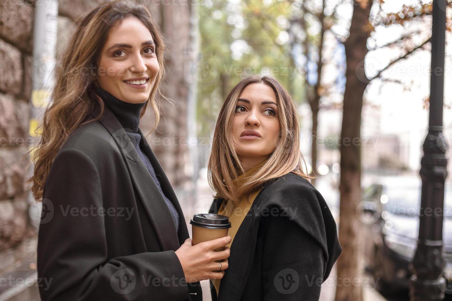 Two happy smiling women female friends with take away coffee standing on city street outdoors photo