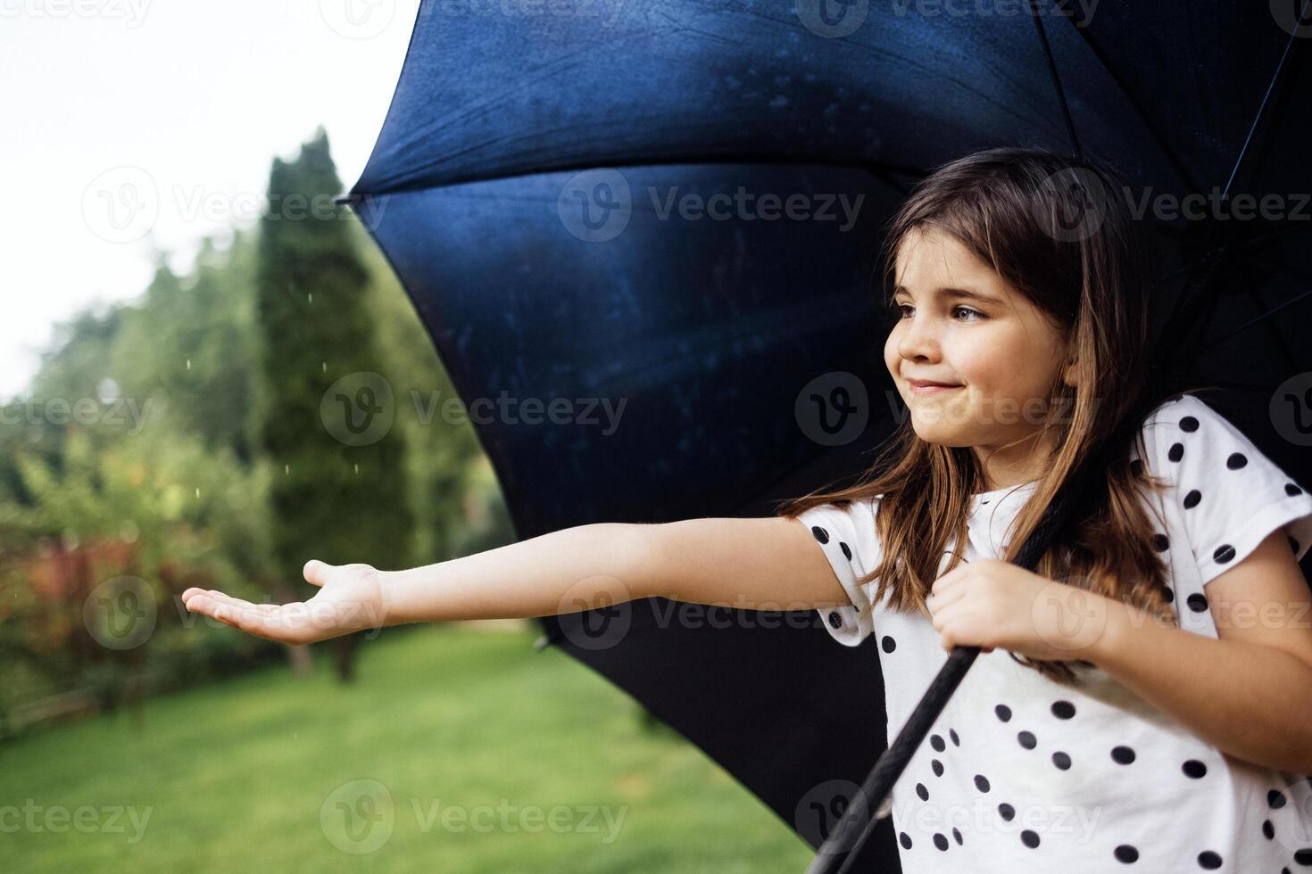 A little girl stands with a black umbrella in the rain. A cute baby stretches out her hand and catches raindrops in the park. photo