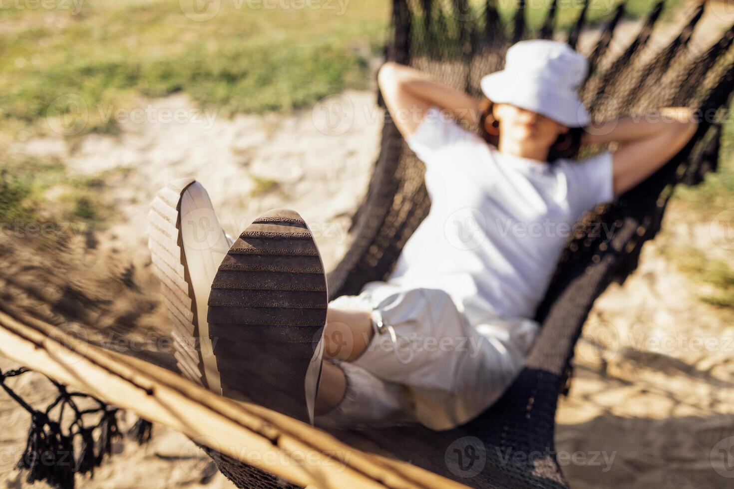 A young woman in casual clothes has her hands behind her head and is lying in a hammock. A girl in a panama hat relaxes and dozes in nature. photo