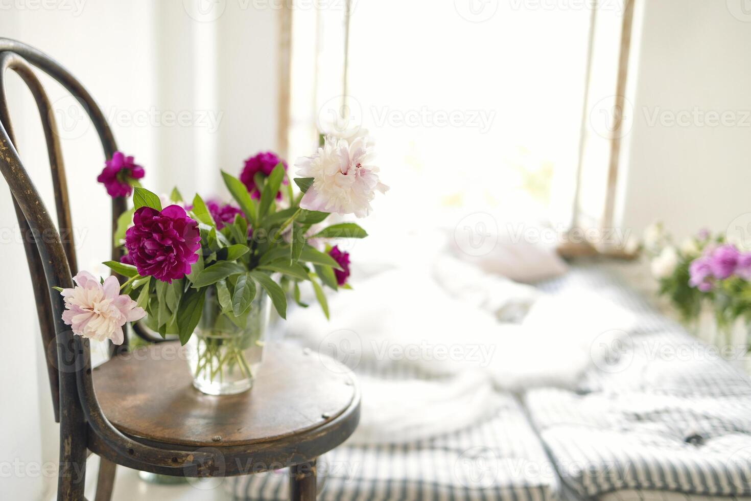 Close up of peonies in a vase on a vintage wooden chair. Rustic style interior with mattresses, pillows and flowers near an open window. photo
