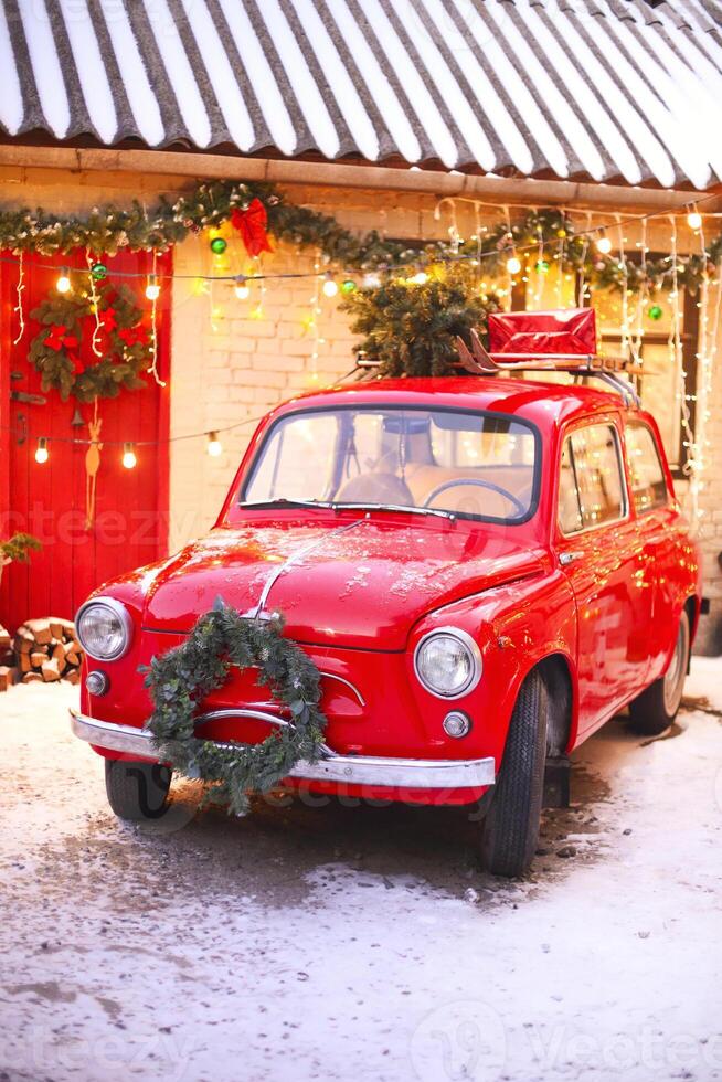 Red retro car in snow-covered courtyard of house decorated with pine branches and xmas lights. photo
