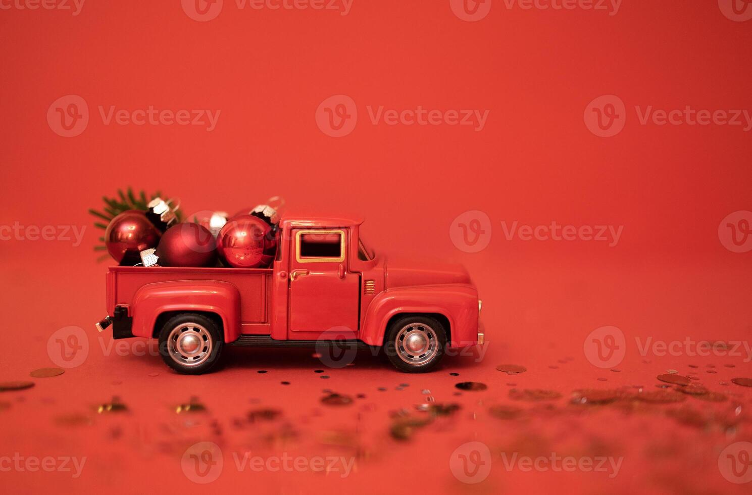 Red toy truck with Christmas balls and fir branch in its trunk. Miniature car with Christmas decoration on red background. Bright backgdrop with sparkles and confetti. photo