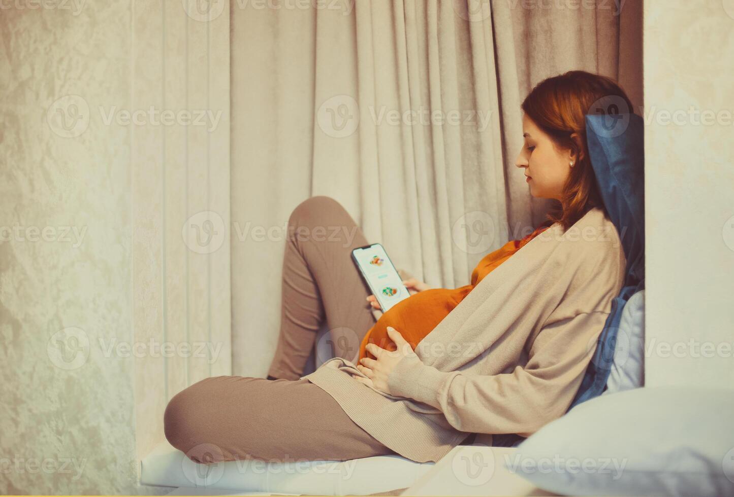 Pregnant woman holding smartphone using pregnancy tracker app on mobile phone while sitting indoors photo