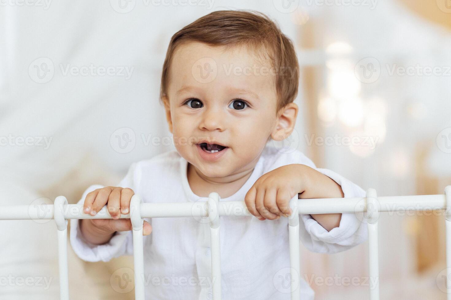 Close-up of a child portrait of a little boy at home in the bedroom. The sweet baby is smiling and holding onto the side of the bed. photo
