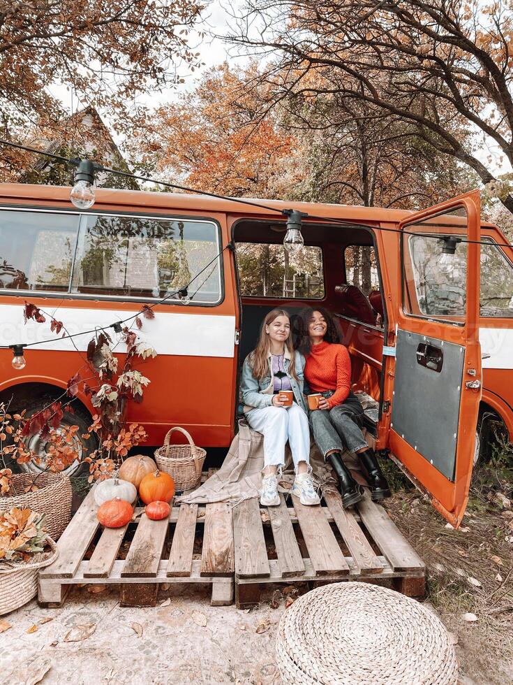 Picnic in autumn nature. Two happy multiracial girlfriends sitting on car trunk in forest photo