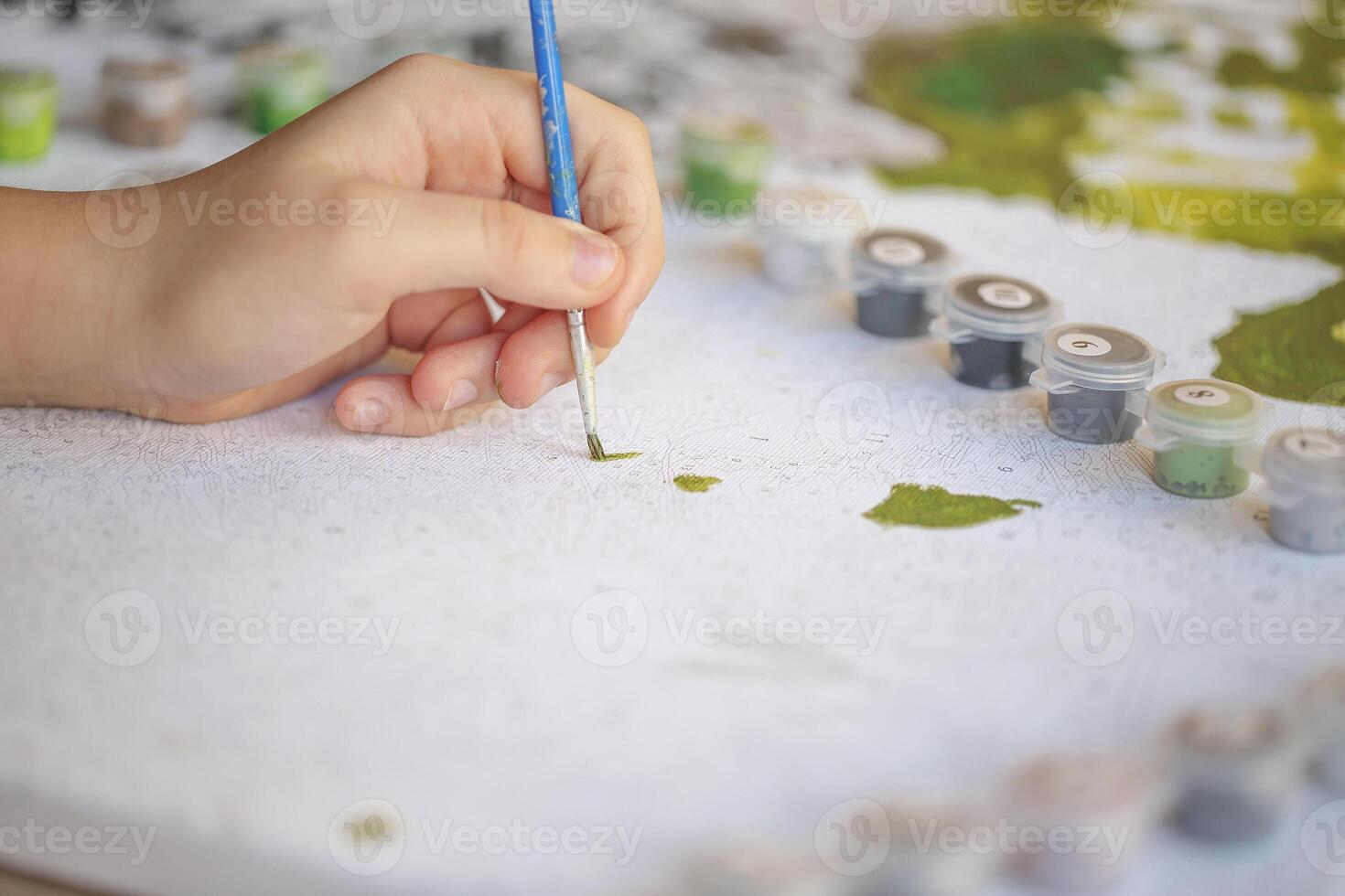 Blurry cropped photo of female hand painting with brush on paper, selective focus on acrylic paints in plastic boxes.