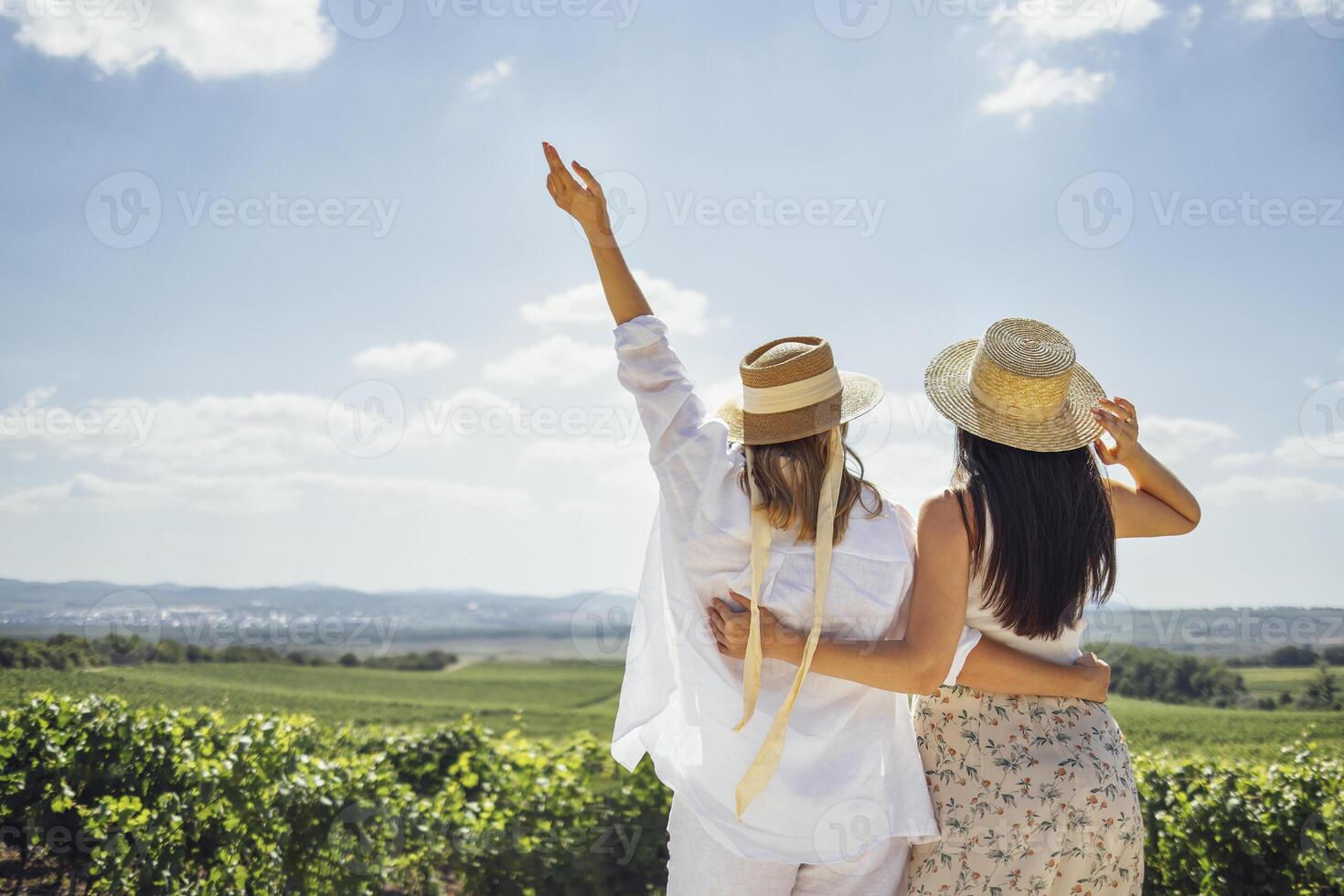 Two happy young women embrace and walk through the vineyard. White female friends in casual clothes and straw hats look at the landscape. photo