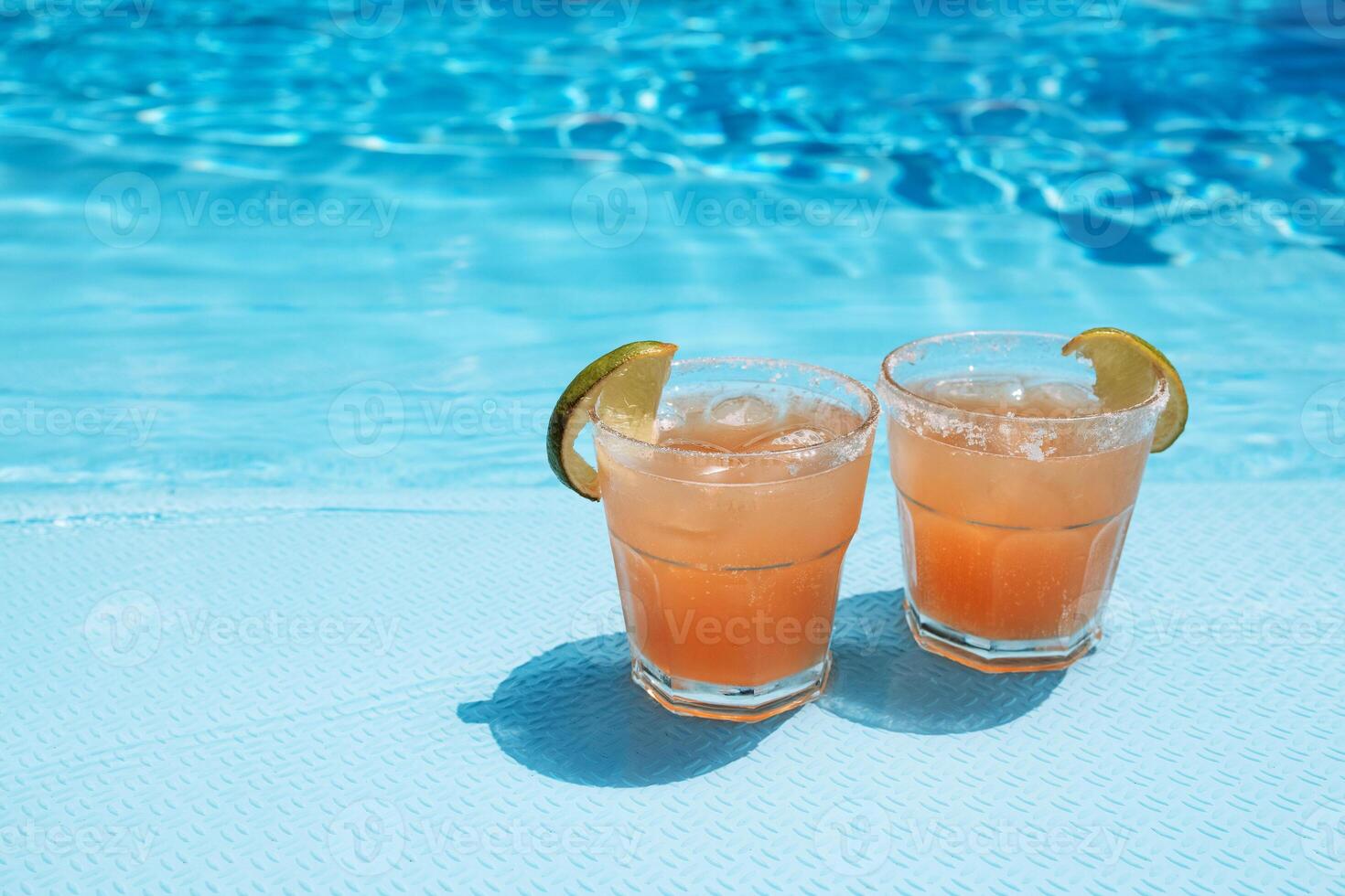 Two glasses with a tasty cocktail with ice cubes. Delicious fresh drink with lime slices by the pool. A sunny day and the reflection of rays in the blue water in the background. photo