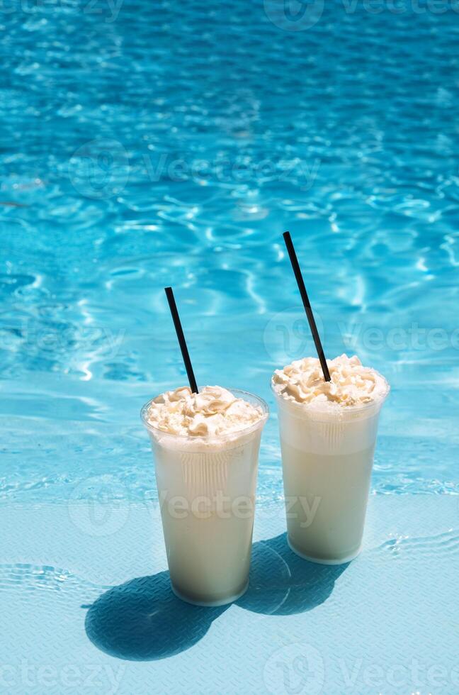 Two glasses with a tasty chilled cocktail. Milkshake with whipped cream. Delicious fresh drink by the pool. A sunny day and the reflection of rays in the blue water in the background. photo