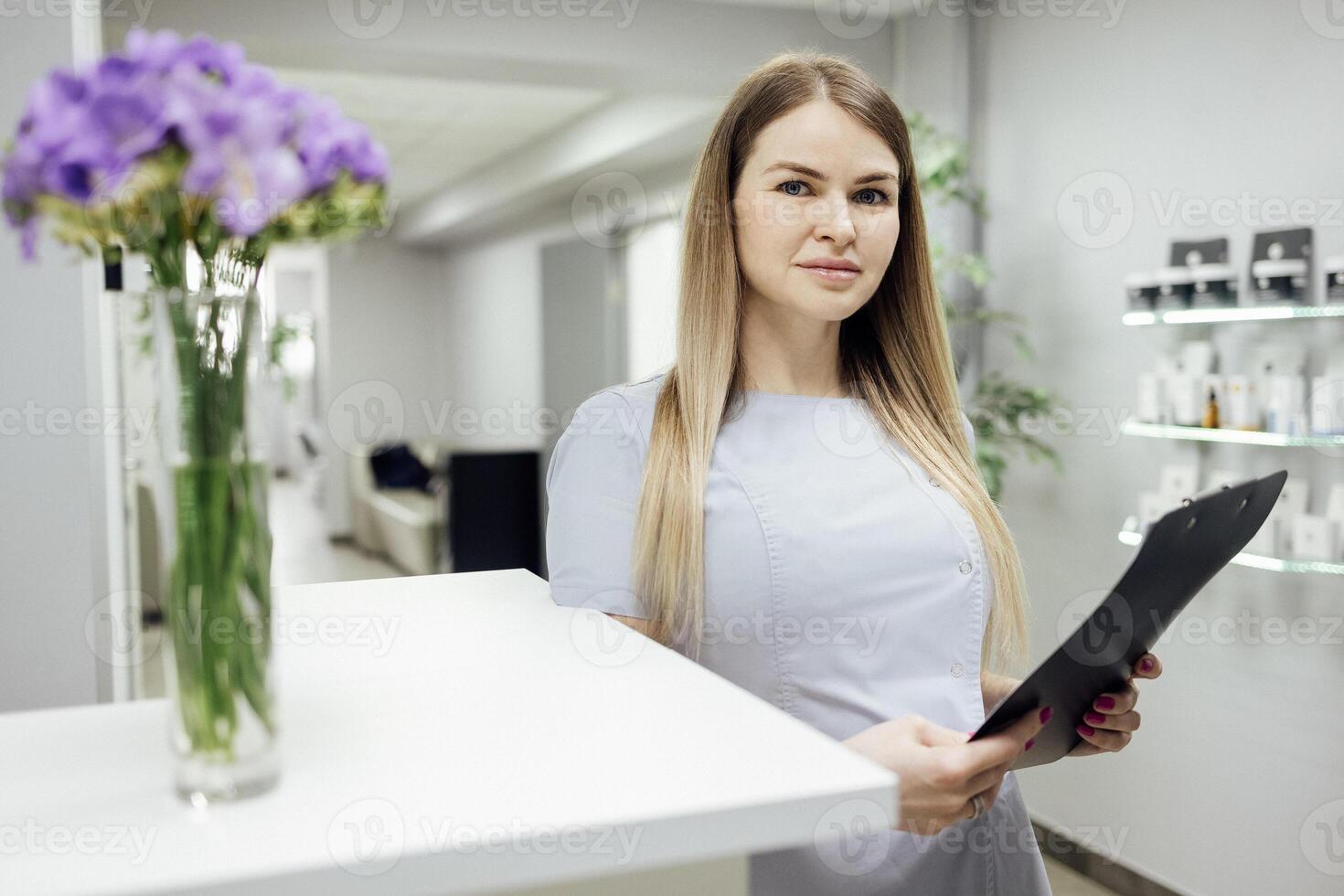 Beautiful female blonde with paper forms at the reception. Young woman in light uniform meets at the counter. Modern interior with beauty product mockups and other rooms on background. photo