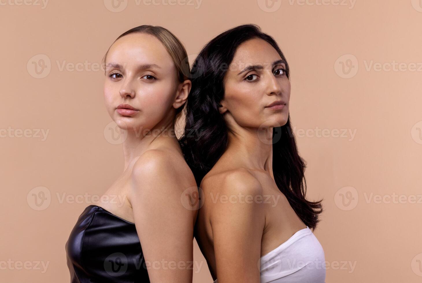 Two young diverse women wearing tops looking confident at camera while posing together on beige background. Charming models with natural makeup and perfect skin. photo