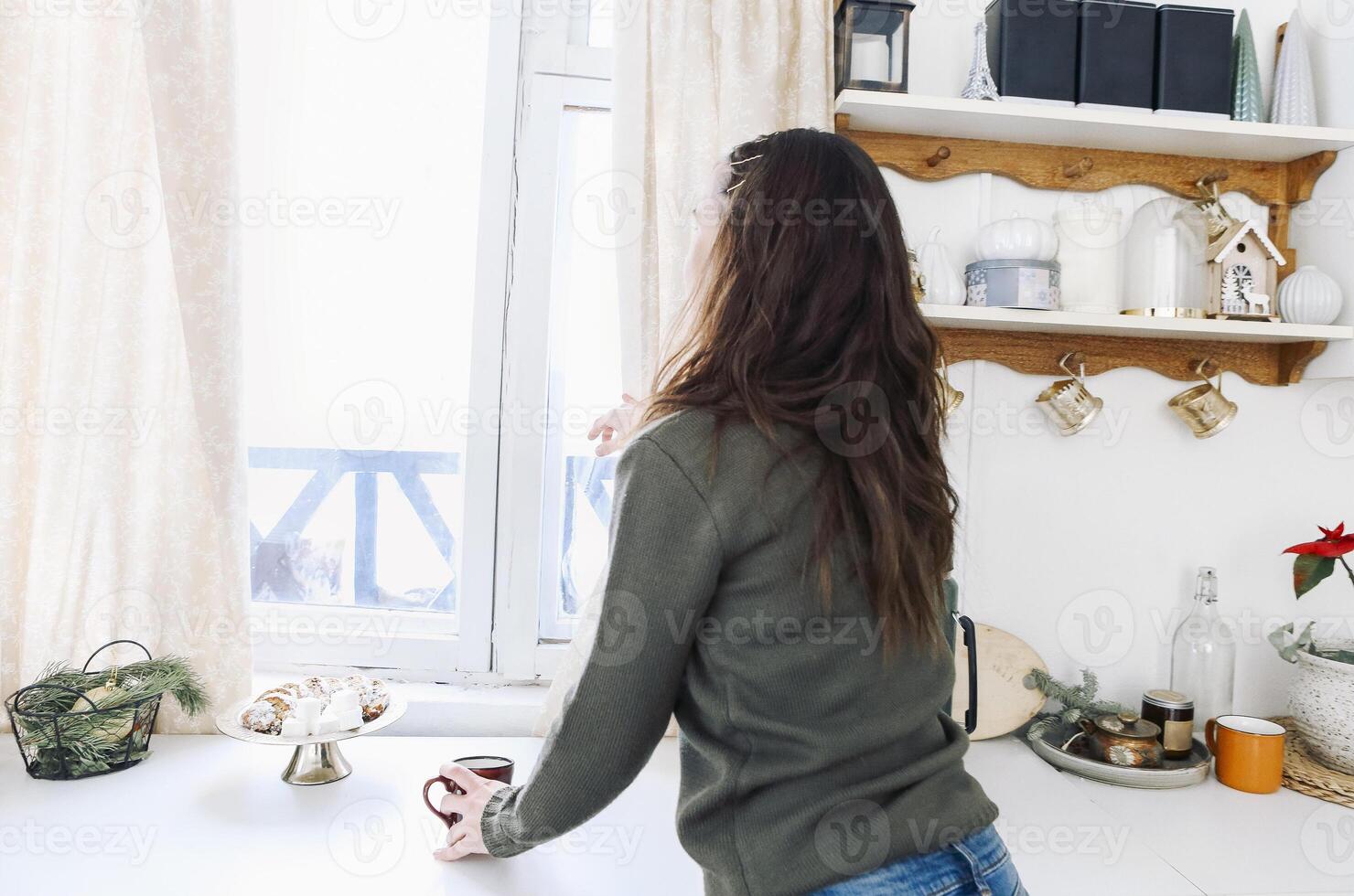Happy woman looking out of kitchen window and drinking hot tea, enjoying New Year holidays at home. photo