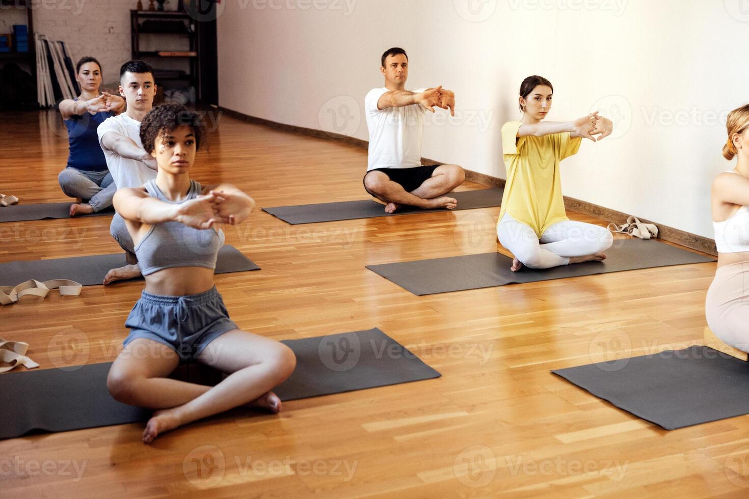 Athletic people of different ages and races practice yoga asanas with trainer. Young multiethnic sporty women and men sit cross-legged on mats, look at coach and stretch out in gym. photo
