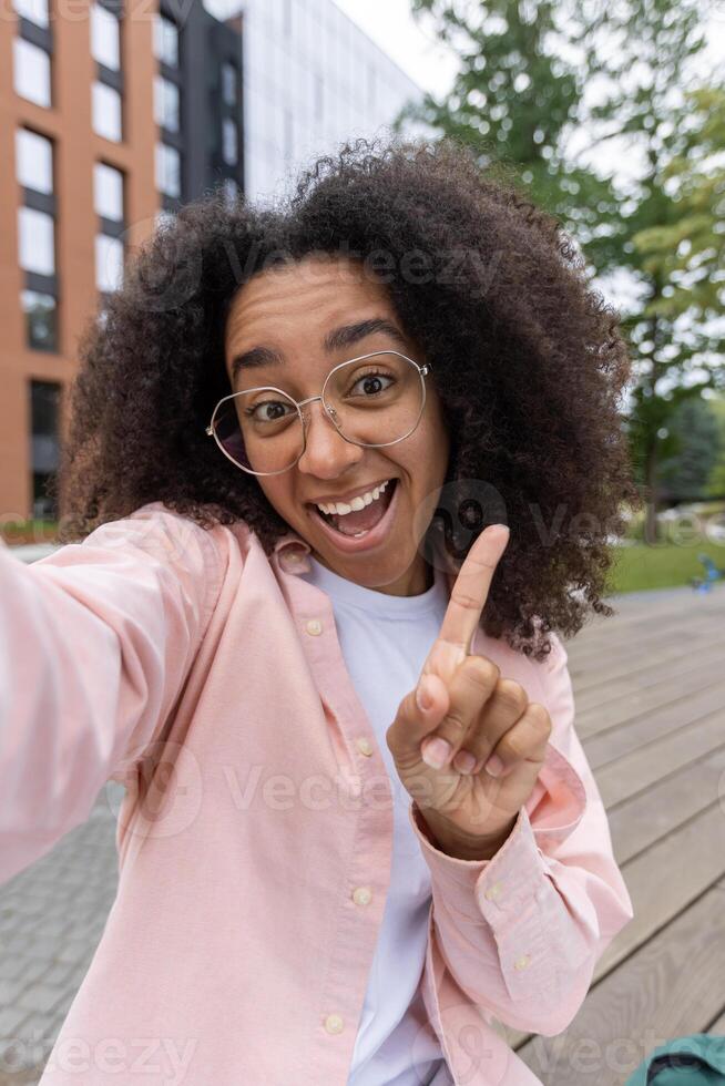 Happy woman smiling and taking a selfie outdoors. She is wearing glasses and a pink shirt, with modern buildings and greenery in the background photo