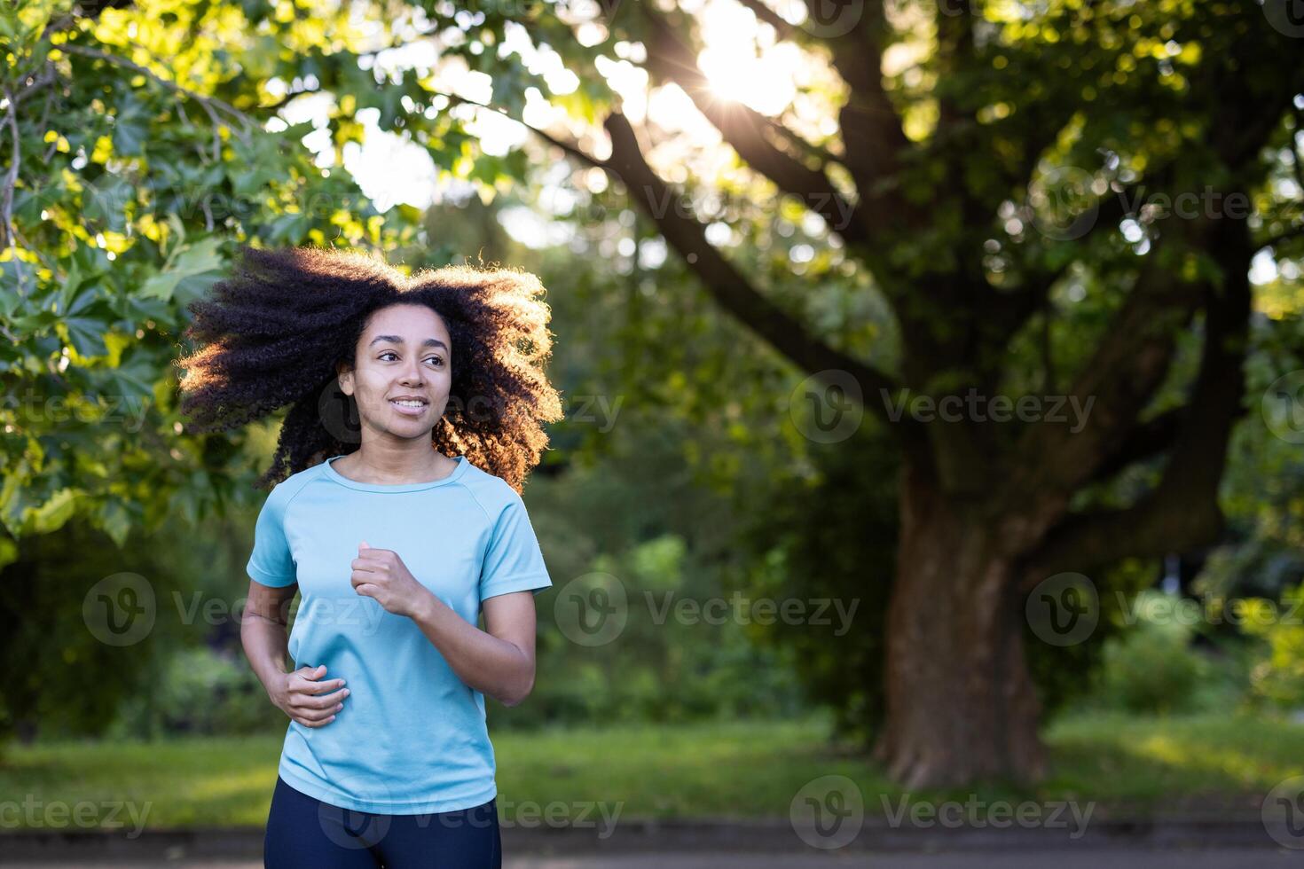 Young African American female athlete jogging outdoors in park. Woman running with sunlight filtering through leaves. Enjoying fitness and healthy lifestyle in natural environment photo