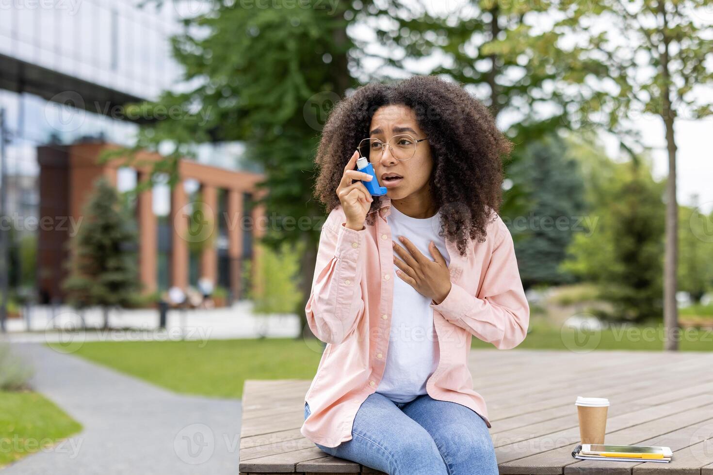 Young woman with asthma using inhaler for relief while sitting on bench in park. She looks anxious and is wearing casual clothes. Green trees and modern buildings in background. photo