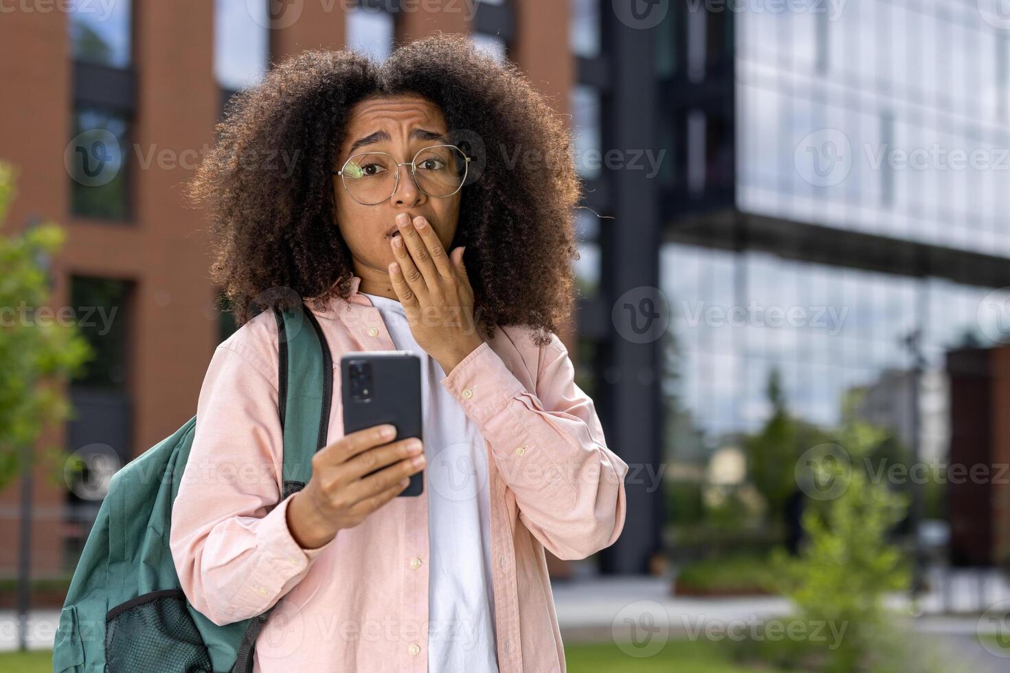 Young student with curly hair and glasses holding smartphone with shocked expression outdoor. Brick and glass building in background. Concept of surprise, technology, urban lifestyle. photo