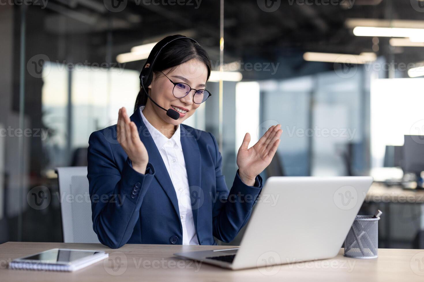 Professional Asian businesswoman in a modern office setting, wearing a headset and conducting a call on her laptop. She appears engaged and confident, sitting at a wooden desk with a notebook photo