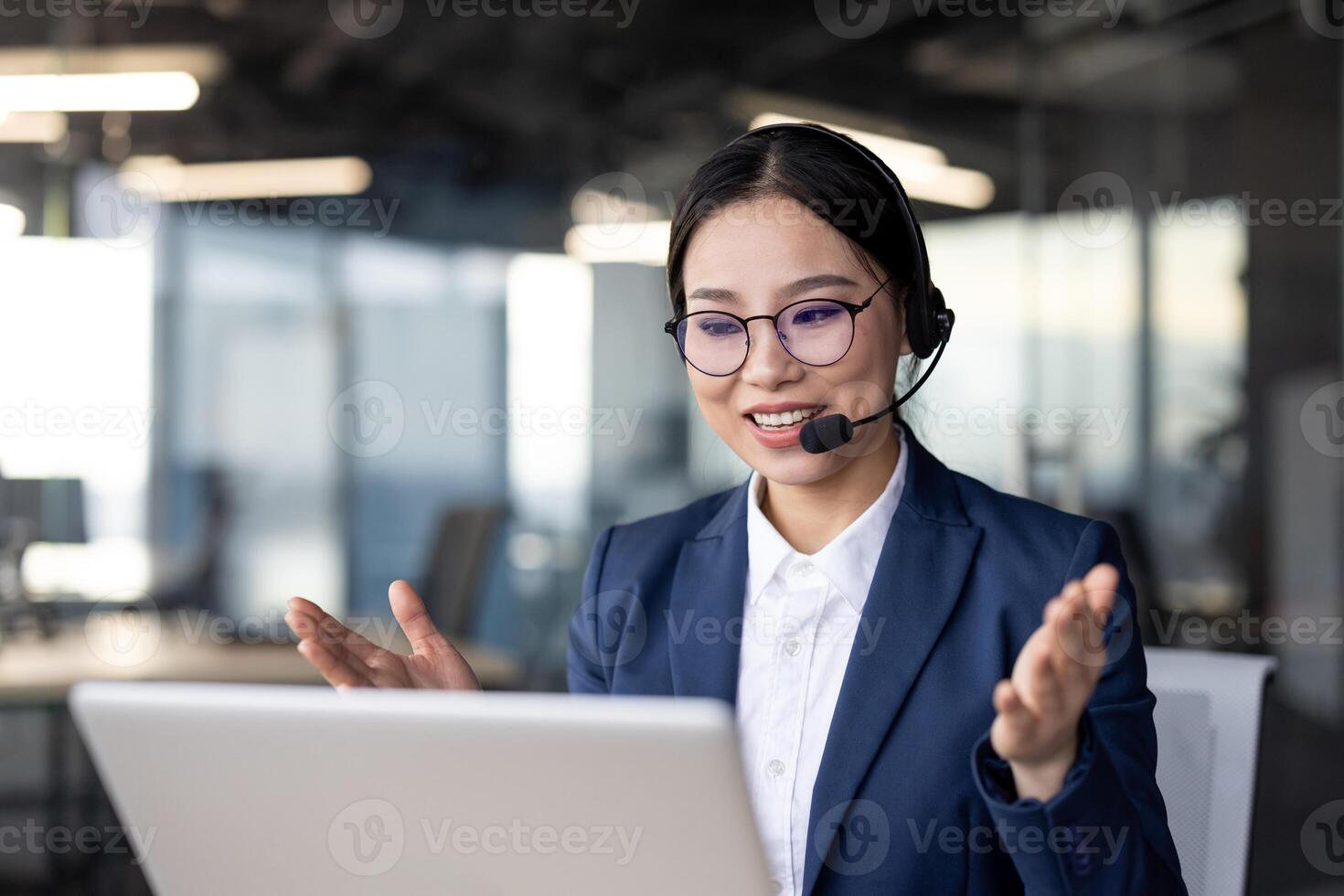 Confident Asian businesswoman wearing a headset and speaking during a virtual meeting. She is seated in a modern office space, actively engaging with her audience while using a laptop. photo