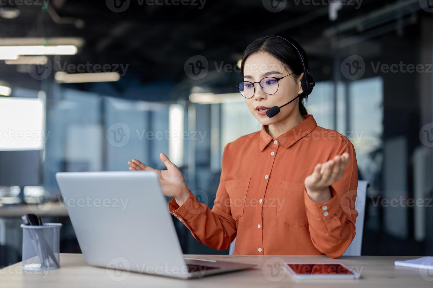 Serious asian woman with headset talking remotely using laptop for call, help desk worker explaining to customers, business woman inside office at work, helpline worker. photo