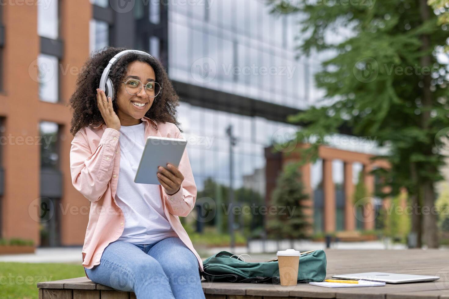 Smiling young woman using digital tablet and headphones while sitting outdoors on university campus. Enjoying music and technology, wearing casual clothes. Relaxed and happy vibes on sunny day. photo