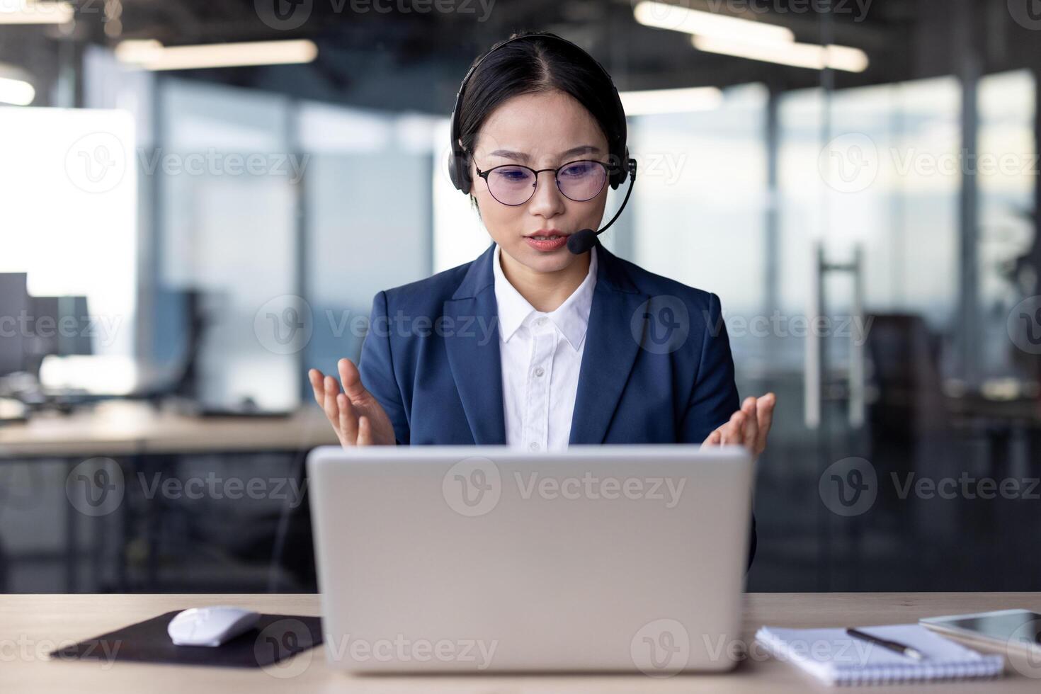 Professional Asian businesswoman in formal attire using a headset for a conference call in a modern office. She appears focused and engaged in a business discussion at her laptop. photo