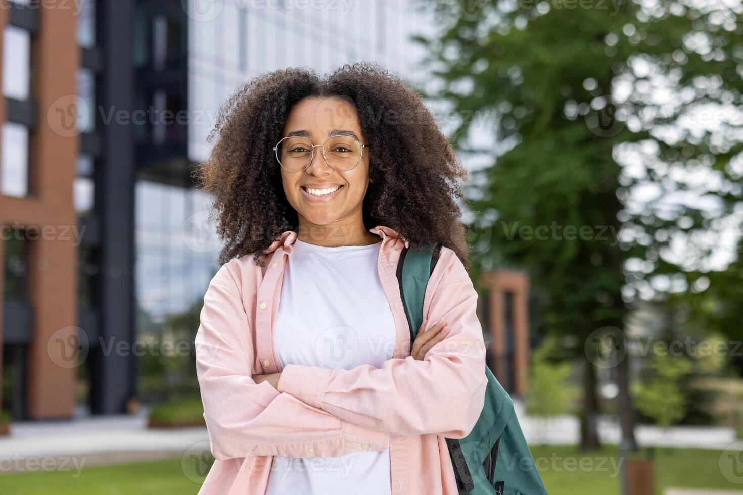 Confident young female student smiling near university campus with backpack. Student wearing glasses and casual clothing standing outside modern building, showing positive and happy demeanor. photo