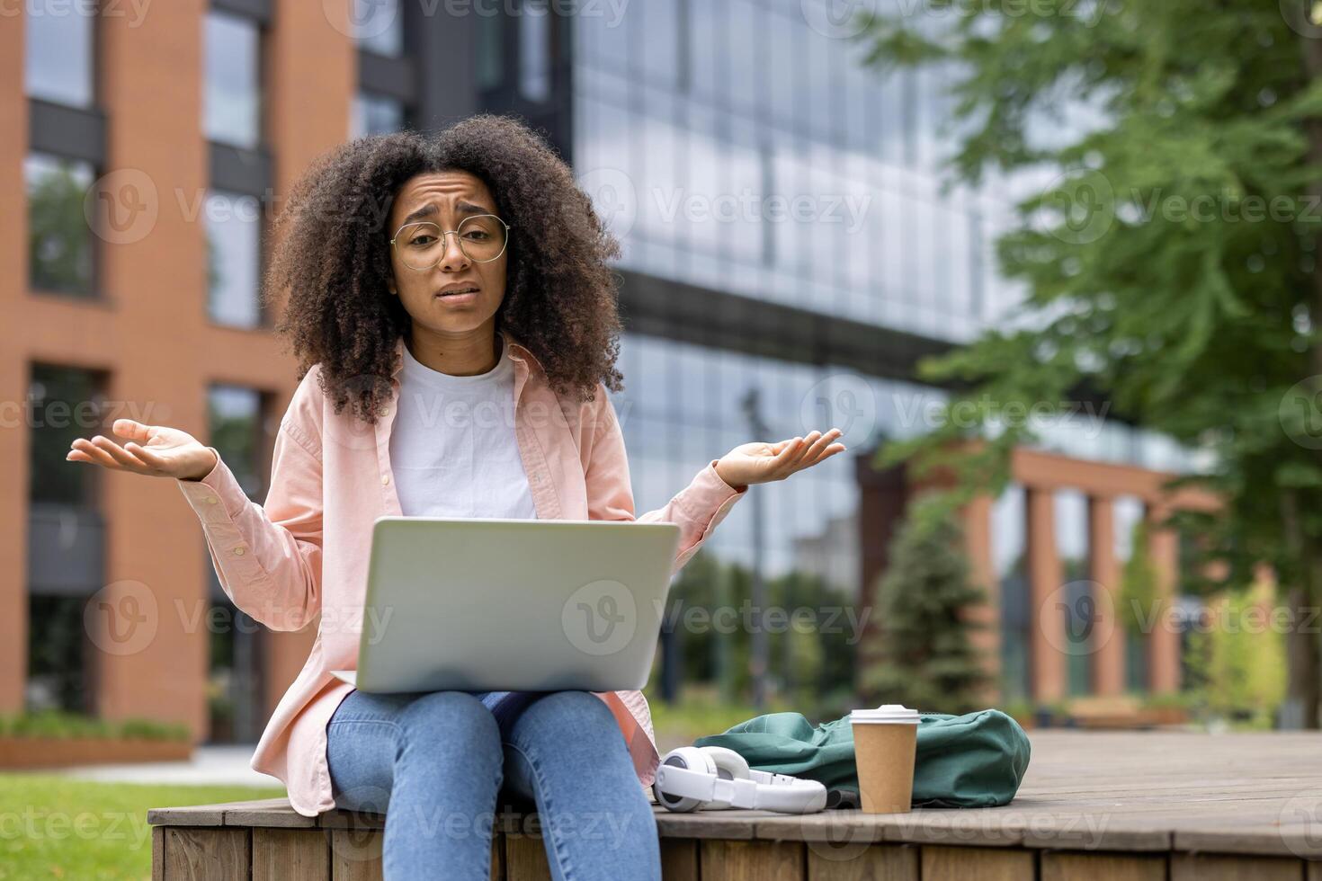 Young woman sitting on bench at university campus using laptop with confused expression. Coffee cup and backpack next to her. Student frustration and outdoor learning concept. photo