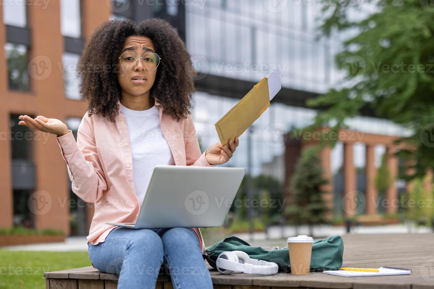 Confused student holding envelope while using laptop. Outdoor workspace in urban setting. Person expressing confusion or misunderstanding. Concept of remote study and modern education methods. photo