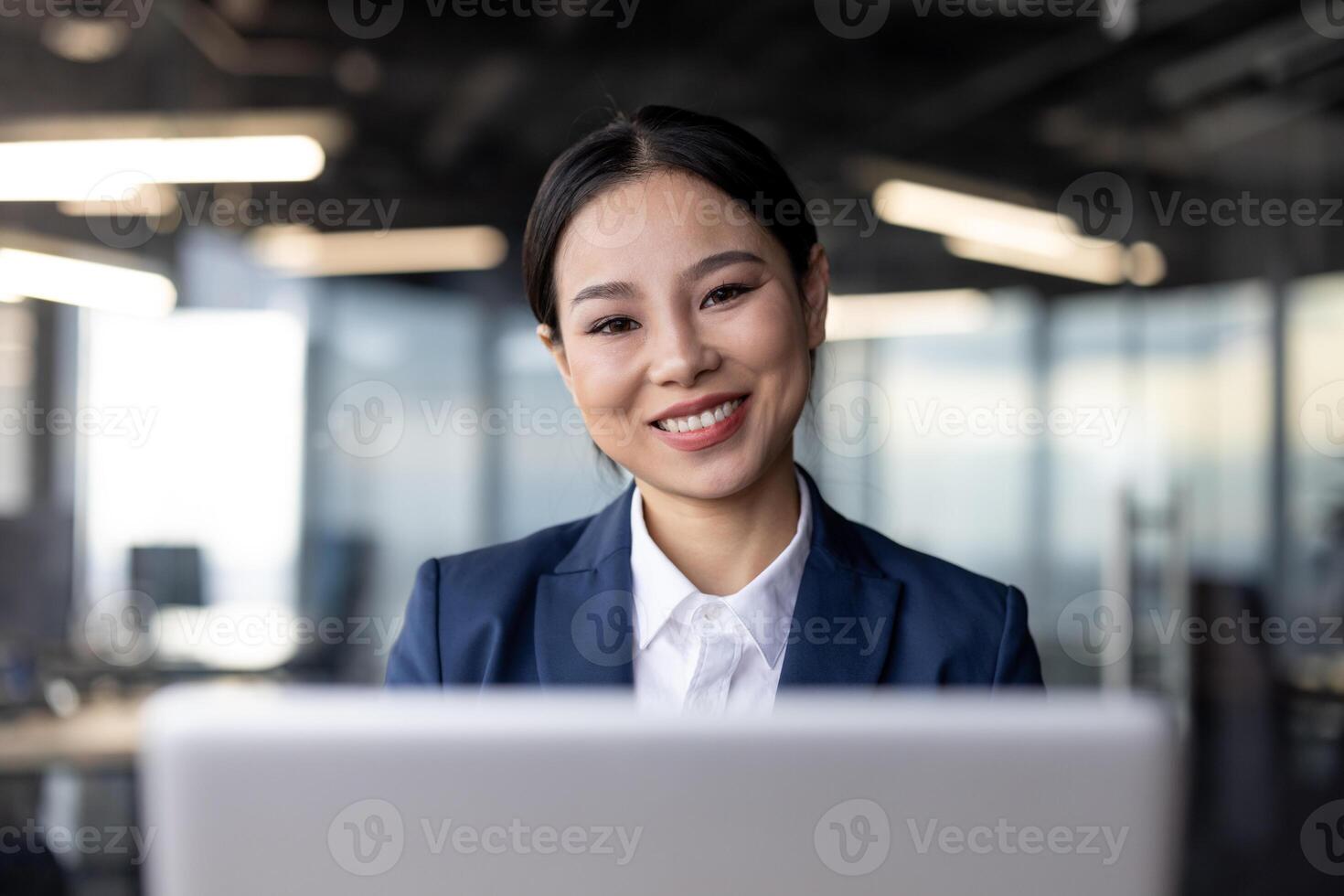Asian businesswoman in formal attire working on a laptop in a modern office setting. She is smiling confidently, embodying professionalism and positivity. photo