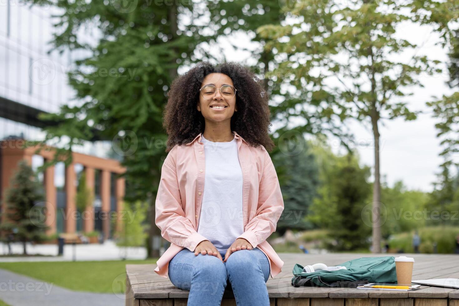 Young woman sitting outdoors smiling, enjoying relaxation and mindfulness. Trees and modern buildings in background. Concepts include relaxation, happiness, nature, and peaceful living. photo