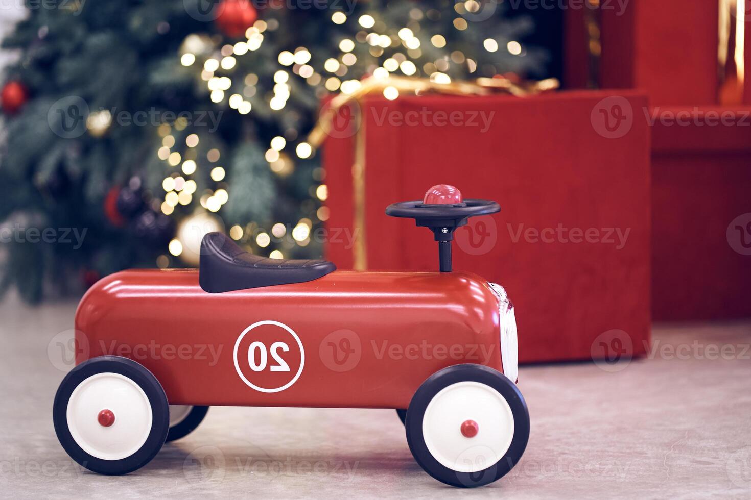 View of red children's retro car on the background of gifts in red boxes with golden bows, Christmas tree decorated garland, string light, red and blue balls. Background, copy space, bokeh photo