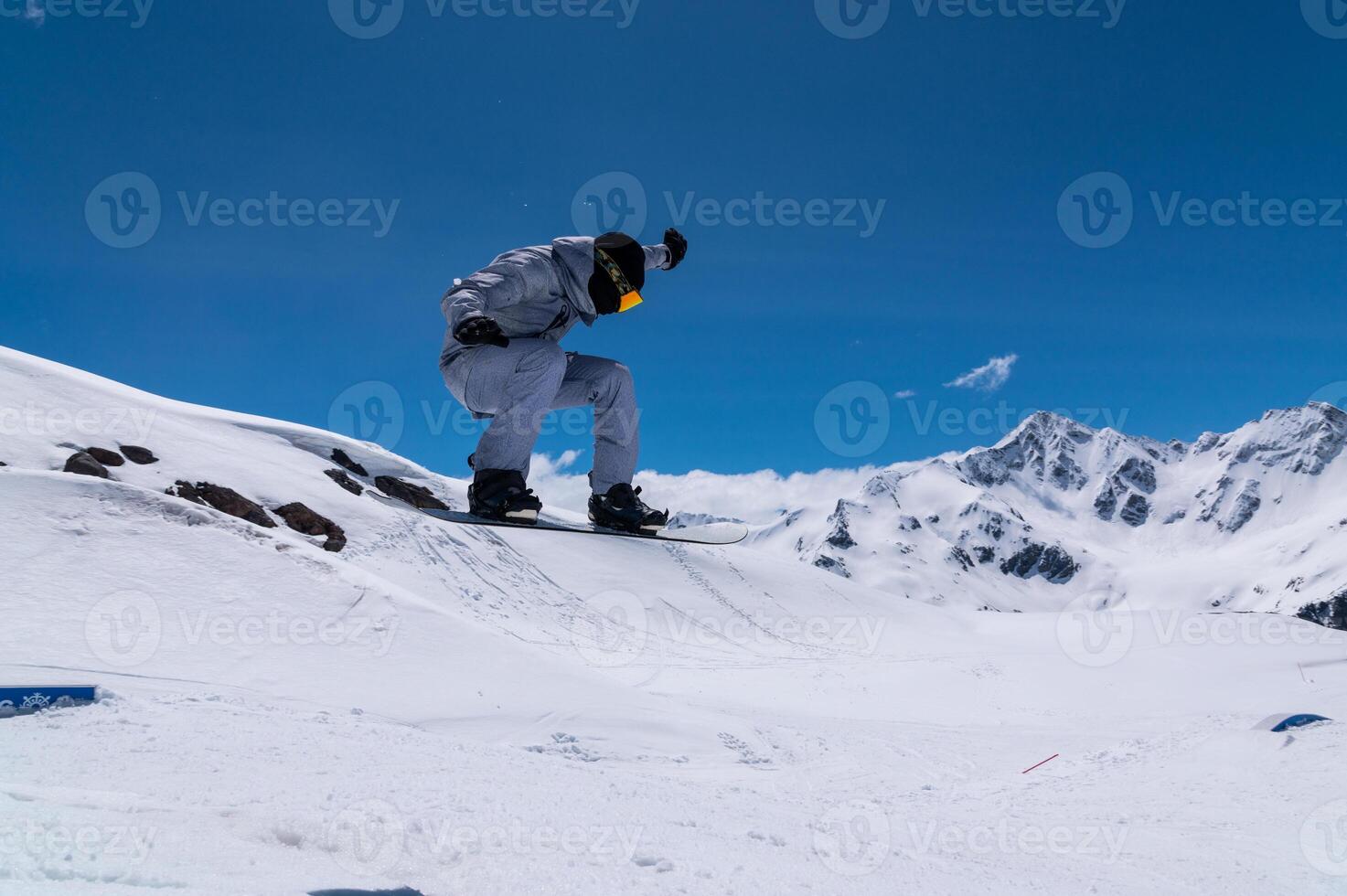 Portrait of a young man snowboarder jumping on a snowboard in sportswear, against the backdrop of high snow-capped mountains and sky photo