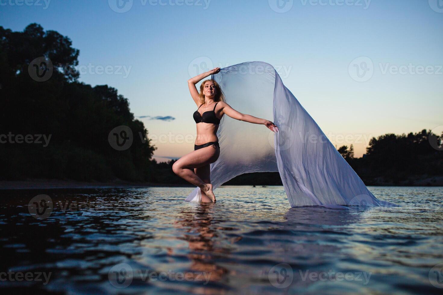 Young slender girl in a black swimsuit posing in the water at sunset with big piece of white fabric photo