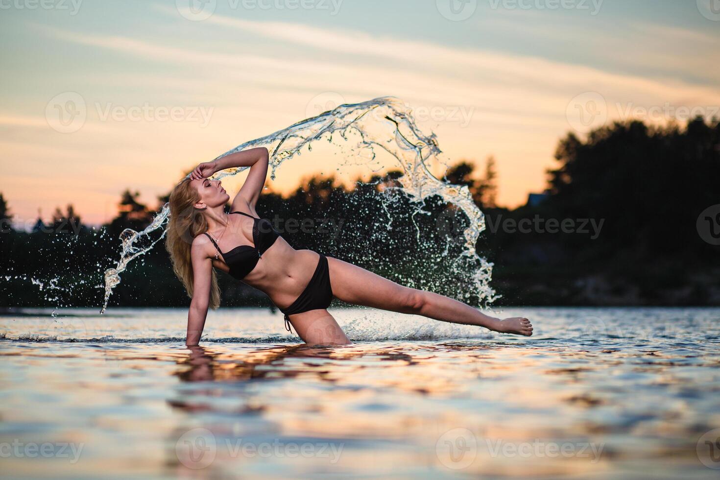 Young slender girl in a black swimsuit posing in the water at sunset at the background of water flying in the air photo