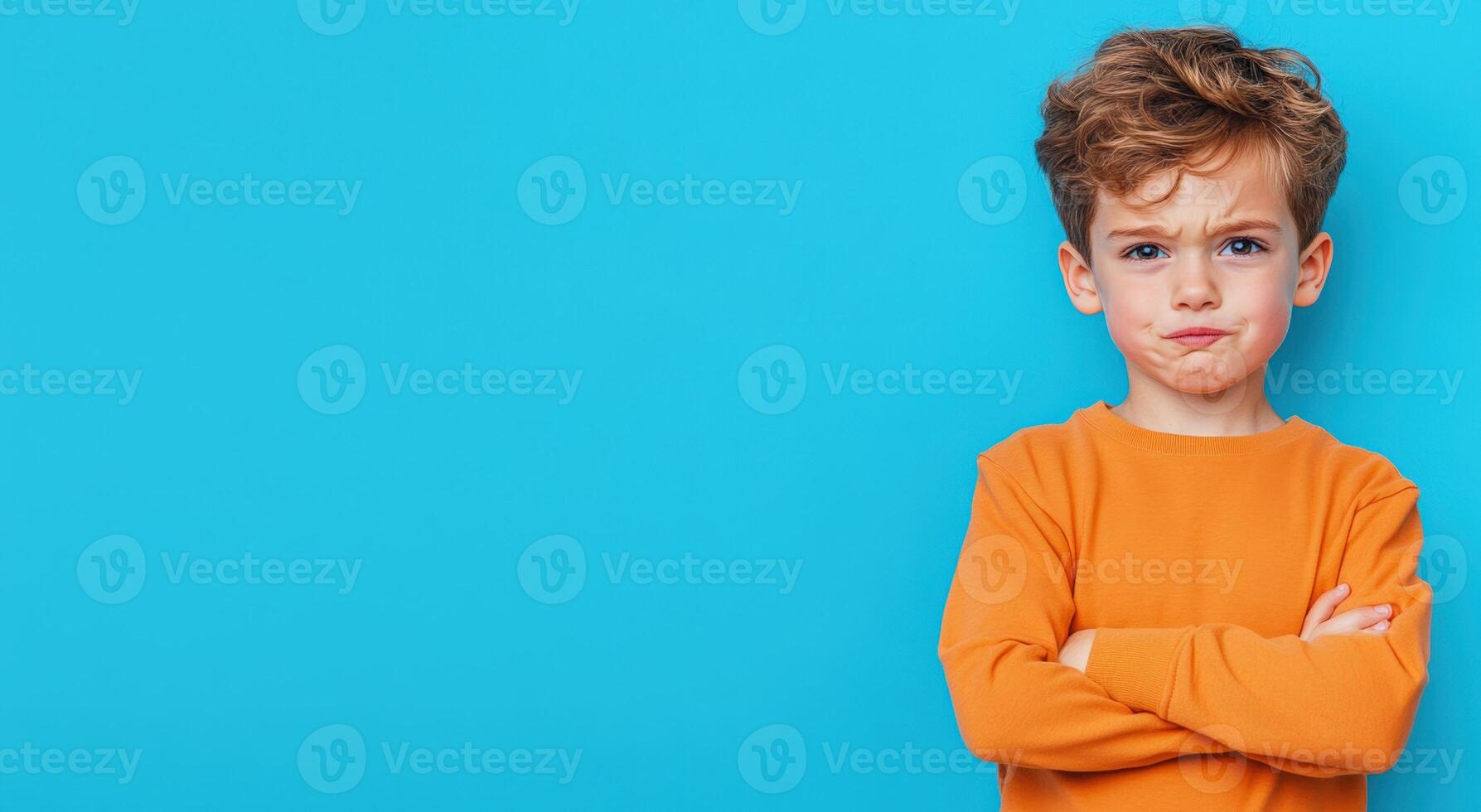 A young boy with curly hair stands with crossed arms, expressing an unhappy look against a bright blue background. photo