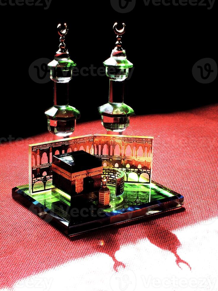 Small model of the Kaaba, the holy mosque of the Grand Mosque, Mecca, display at the top of red fabric material and dark background. There is sunlight coming from behind to form an amazing reflection photo
