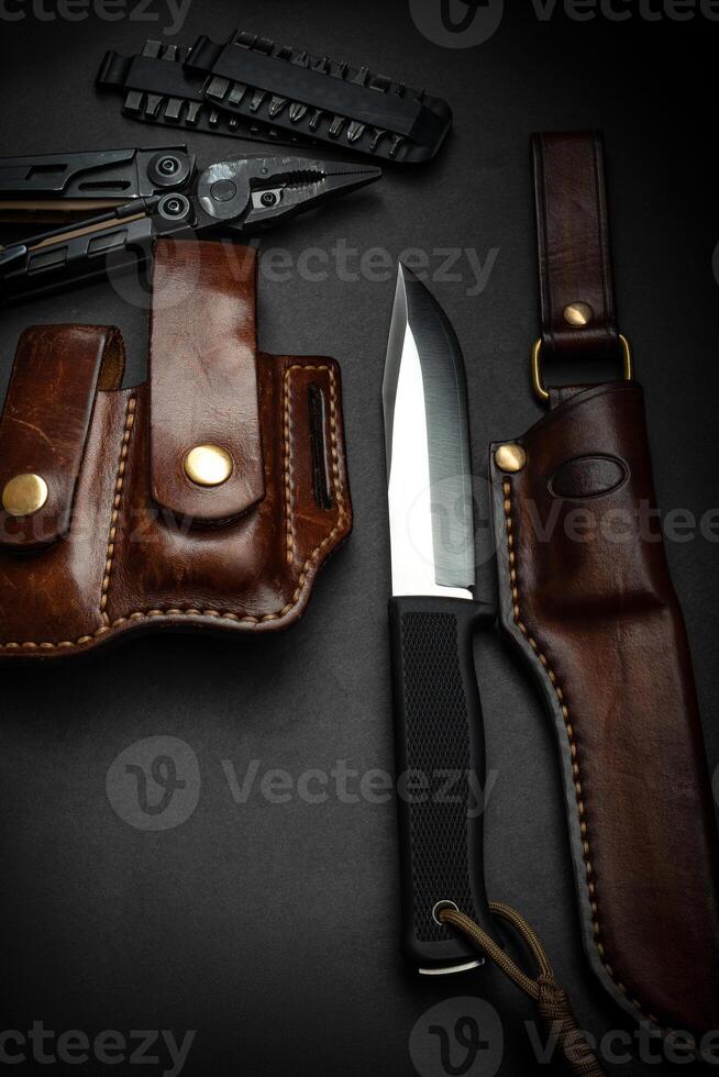 Hunting knife and multitool on a dark background. Brown leather cases near the knife and multitool. Postcard concept for men. photo